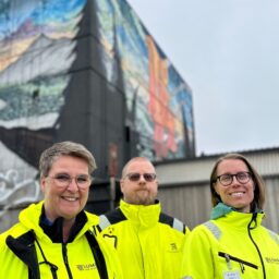 Bild på tre personer utomhus i gula arbetskläder framför industribyggnad med grafittimålning