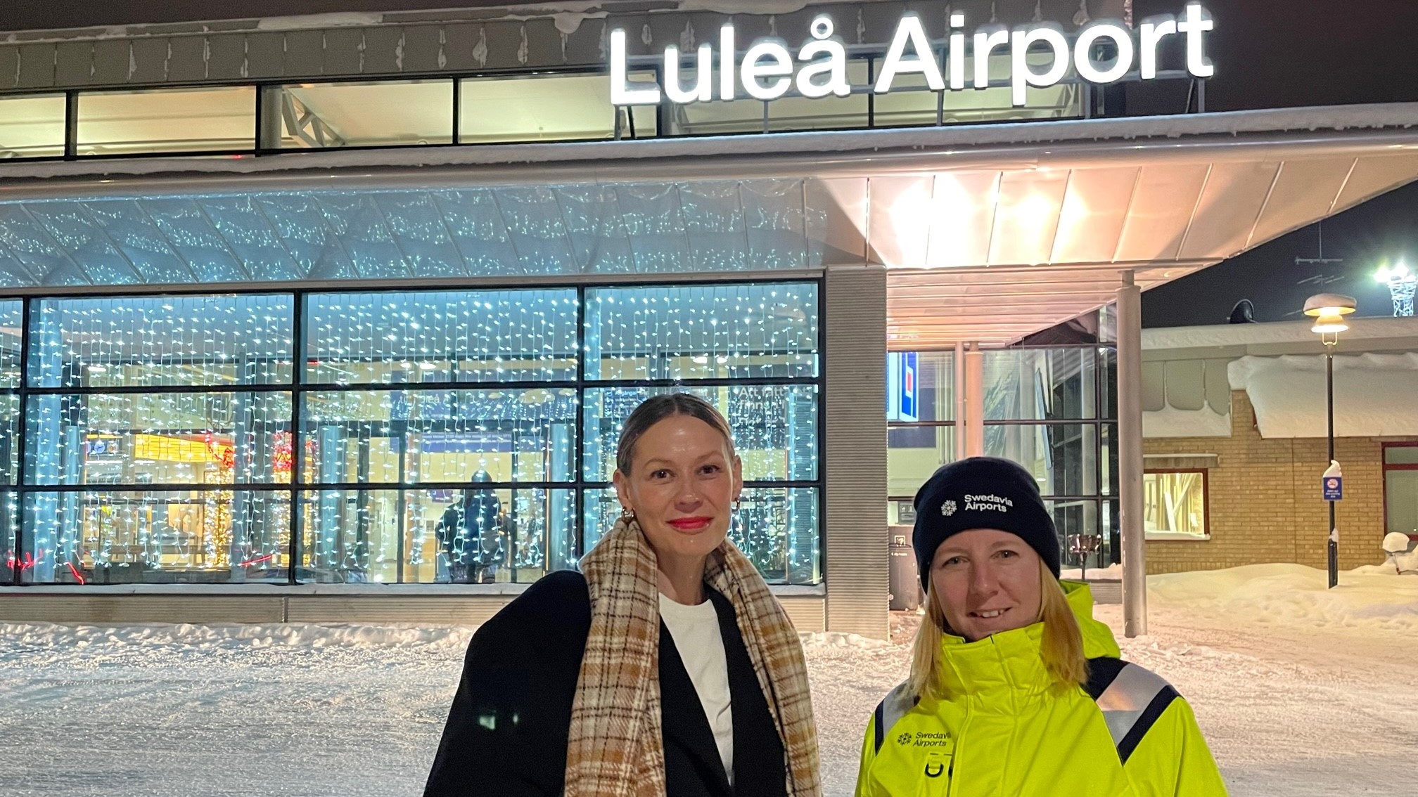 Två personer utomhus i vinterlandskap vid flygplatsskylt
